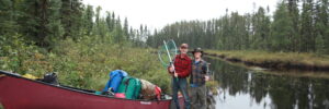 Two people stand by a red canoe on a riverbank, holding fishing nets. The area is surrounded by trees and various camping gear.