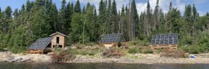 Three small wooden cabins with solar panels are situated by a lake, surrounded by a dense forest under a partly cloudy sky.