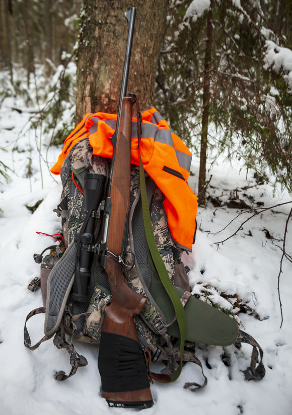 Hunting carbine stands at the trunk of tree next to backpack and red vest after roundup of winter hunting for ungulates. Rifle with an optical sight rests on backpack with signal cape in snowy forest.