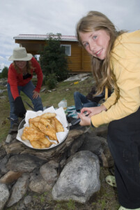 Girl holds a pan with fried fish outdoors, while another person cooks nearby. A log cabin and overcast sky are in the background.